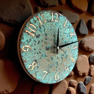 Puede incluir: Reloj de pared redondo con esfera turquesa y marrón, con números y manecillas de color cobre. El reloj está montado en una pared de piedra texturizada, creando una estética rústica. La esfera del reloj tiene un aspecto moteado único.