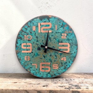 Può includere: Orologio da parete rotondo con finitura turchese e rame. Il quadrante dell'orologio ha un aspetto strutturato con numeri e lancette color rame. L'orologio è esposto su una superficie di legno.