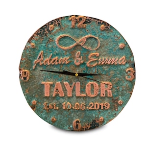 Puede incluir: Un reloj de cobre rústico con una pátina verde desgastada. La esfera del reloj presenta números romanos, un símbolo de infinito y los nombres "Adam & Emma Taylor" con la fecha "Est. 10.06.2019".
