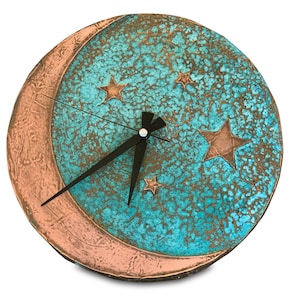 Puede incluir: Un reloj redondo de color turquesa y cobre con un diseño de luna creciente y estrellas. El reloj tiene agujas negras y una esfera blanca.
