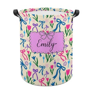 May include: A cylindrical laundry hamper with a floral pattern of pink tulips, blue bows, and green leaves on a cream background. A pink label with the name "Emily" is centered on the front. Black handles are attached to the top.