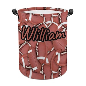 May include: A cylindrical laundry hamper featuring a repeating pattern of red and white footballs. The name "William" is printed in a black script across the front. The hamper has black handles.
