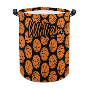 May include: A cylindrical laundry basket featuring a black backdrop adorned with a repeating pattern of orange basketballs. The name "William" is written in a cursive font. It has black handles.