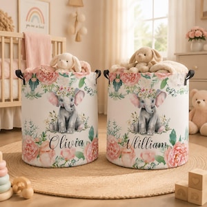 Puede incluir: Dos cestas de almacenamiento cilíndricas de tela con un estampado de acuarela de un elefante gris con una corona de flores, rodeado de flores rosas y melocotón y follaje verde. Una cesta está personalizada con el nombre Olivia y la otra con William.