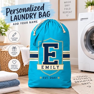May include: A bright blue laundry bag with a drawstring closure, featuring a large navy blue letter E inside a gold shield emblem. Below the shield, the name EMILY is printed in navy blue on a gold banner, with the text EST. 2027 and two gold stars.