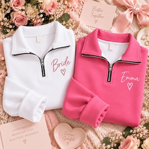 May include: A white quarter-zip sweatshirt with the word Bride and a heart printed in dark pink, and a hot pink quarter-zip sweatshirt with the name Emma and a heart printed in white, both featuring black zippers and fold-over collars.