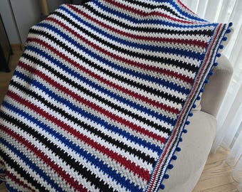 Puede incluir: Una manta de crochet a rayas con un esquema de colores azul, rojo, blanco, negro y gris. La manta tiene un borde de crochet con pompones azules.