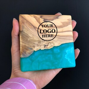 May include: A square wooden coaster with a turquoise blue epoxy resin inlay. The wood has a natural grain pattern and the resin has a swirling effect. The coaster has the text "YOUR LOGO HERE" engraved in the wood.