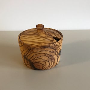 May include: A small, round wooden salt or sugar dispenser with a lid. The dispenser is made of olive wood and has a natural wood finish. The lid has a small hole for pouring.