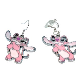 Puede incluir: Un par de pendientes de esmalte rosa y blanco con un dise&ntilde;o de personaje de dibujos animados. Un pendiente tiene un gancho, el otro un cierre de clip. Los pendientes tienen un borde de metal plateado.