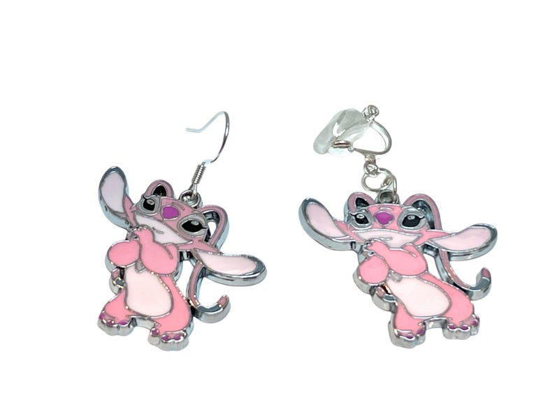 Puede incluir: Un par de pendientes de esmalte rosa y blanco con un dise&ntilde;o de personaje de dibujos animados. Un pendiente tiene un gancho, el otro un cierre de clip. Los pendientes tienen un borde de metal plateado.