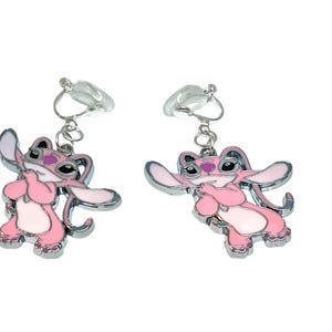 Puede incluir: Un par de pendientes de clip con un personaje de dibujos animados rosa con ribetes plateados. El personaje tiene orejas grandes, una nariz peque&ntilde;a y se sujeta las manos delante del pecho. Los pendientes est&aacute;n sujetos a clips de pl&aacute;stico transparente.