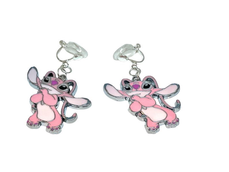 Puede incluir: Un par de pendientes de clip con un personaje de dibujos animados rosa con ribetes plateados. El personaje tiene orejas grandes, una nariz peque&ntilde;a y se sujeta las manos delante del pecho. Los pendientes est&aacute;n sujetos a clips de pl&aacute;stico transparente.
