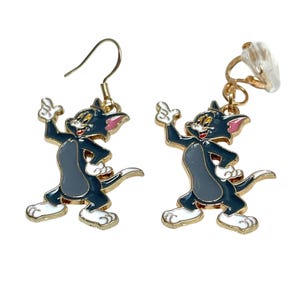 May include: A pair of Tom and Jerry cartoon character earrings. The earrings feature a gold-colored metal frame with a cartoon cat in a standing pose. The cat is primarily gray with white and pink accents.