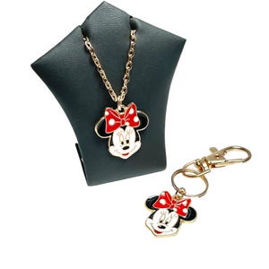 May include: A gold-toned necklace and keychain set featuring a Minnie Mouse design. The pendant and keychain charm have a red bow, black ears, and a white face. The necklace is displayed on a dark green stand.