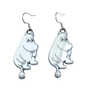 以下が含まれることがあります： 銀色のフックが付いた、白いムーミンのキャラクターのイヤリングのペア。各イヤリングは、笑顔の顔、腕、脚、尻尾を持つ漫画のキャラクターが特徴です。イヤリングは白いエナメル仕上げと銀色の輪郭でデザインされています。