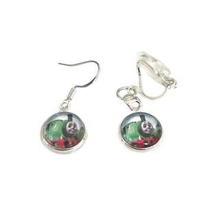 May include: A pair of silver-toned earrings featuring a circular image of a green train with a smiling face, set against a red and green background. One earring has a hook, the other a clip-on style.