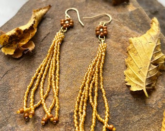 May include: Gold-coloured beaded earrings featuring a floral design at the top and multiple bead strands. The earrings are presented on a textured, brown surface with dried leaves.