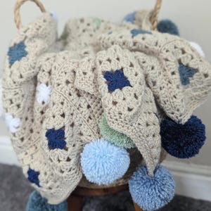 May include: A beige crocheted blanket with blue, white, and green square accents and blue pom-poms. The blanket is draped over a small wooden stool. The blanket has a basket-weave design and is handmade.