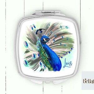 Peut inclure: Un miroir de poche avec un cadre argent&eacute; et un fond blanc. Le miroir pr&eacute;sente une illustration color&eacute;e d'un paon avec ses plumes de queue d&eacute;ploy&eacute;es. Le nom "Lois" est &eacute;crit en cursive sous le paon.