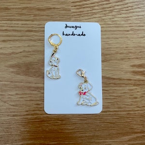 Peut inclure: Deux breloques de chien dalmatien dorées avec des nœuds rouges. Les breloques sont attachées à des porte-clés. Les breloques sont sur une carte blanche avec le texte "Juwagi handmade".
