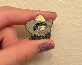 May include: A silver enamel pin featuring a black cat wearing a yellow cowboy hat. The cat has a white beard and a serious expression.