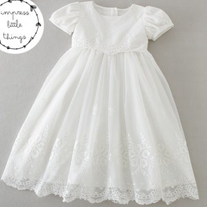 May include: A white, short-sleeved dress with intricate floral embroidery. The dress features a round neckline and a flowing skirt. The image includes the text "impress little things" in a circular design.