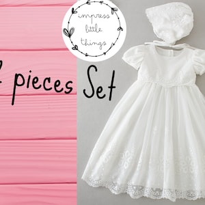 May include: A two-piece white baby dress set. The set includes a short-sleeved dress with floral embroidery and a matching bonnet. The dress has a tulle overlay and lace trim. The text "2 pieces Set" is visible in the image.