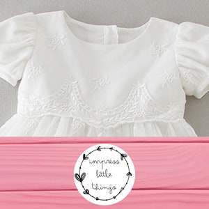 May include: White baby dress with short, puffed sleeves and delicate floral embroidery. The dress features a detailed bodice and a flowing skirt. The image includes a logo that says "impress little things".