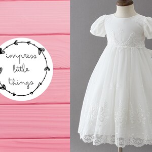 May include: A white formal dress with short, puffy sleeves and a detailed lace bodice and hem. The dress features a delicate crown-like design at the waist. The image also includes a logo that says "impress little things" in a circle.
