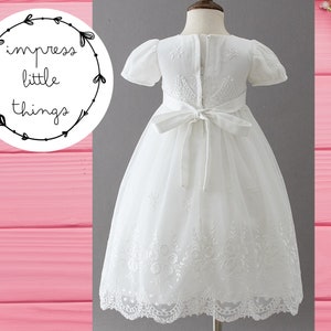 May include: A white dress with short sleeves and a tulle skirt. The dress features embroidered details and a bow at the back. The text "impress little things" is in a circular design in the upper left corner.