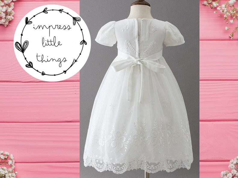 May include: A white dress with short sleeves and a tulle skirt. The dress features embroidered details and a bow at the back. The text "impress little things" is in a circular design in the upper left corner.