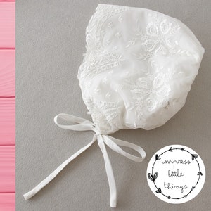 May include: A white, lacy bonnet with intricate floral embroidery and a delicate tie closure. The bonnet is made of sheer fabric and is likely for a baby or young child. The image includes a logo that says "impress little things."