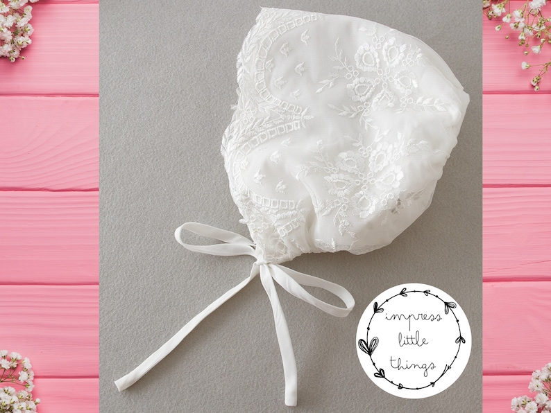 May include: A white, lacy bonnet with intricate floral embroidery and a delicate tie closure. The bonnet is made of sheer fabric and is likely for a baby or young child. The image includes a logo that says "impress little things."