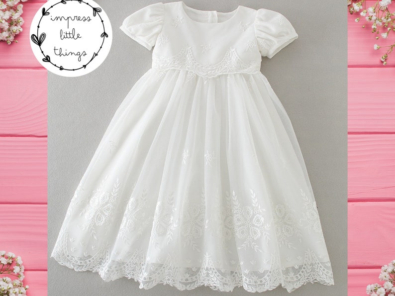 May include: A white, short-sleeved dress with intricate floral embroidery. The dress features a round neckline and a flowing skirt. The image includes the text "impress little things" in a circular design.