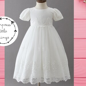 May include: A white formal dress with short, puffed sleeves and a full skirt. The dress features intricate embroidered details on the bodice and hem. The text "impress little things" is in a circular logo on the left.