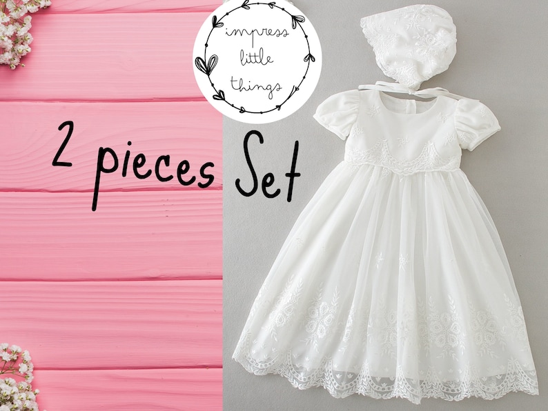 May include: A two-piece white baby dress set. The set includes a short-sleeved dress with floral embroidery and a matching bonnet. The dress has a tulle overlay and lace trim. The text "2 pieces Set" is visible in the image.