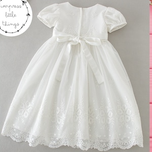 May include: A white, short-sleeved dress with floral embroidery and a bow at the waist. The dress has a layered skirt and is likely for a special occasion. The dress is from "impress little things".
