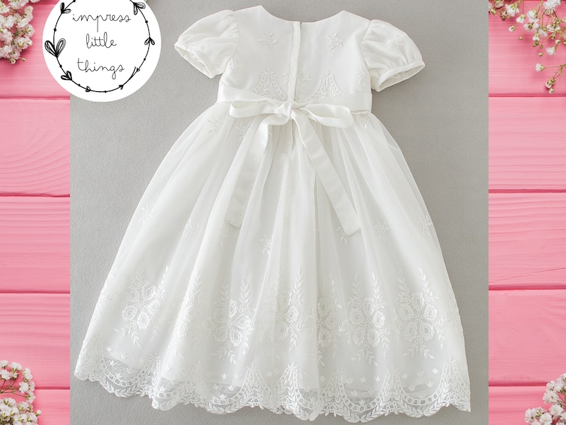 May include: A white, short-sleeved dress with floral embroidery and a bow at the waist. The dress has a layered skirt and is likely for a special occasion. The dress is from "impress little things".