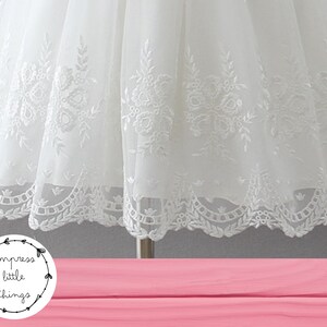 May include: Close-up of a white dress with intricate floral embroidery. The dress features a delicate, layered design with detailed patterns along the hemline. The image includes the text "impress little things" in a circular logo.
