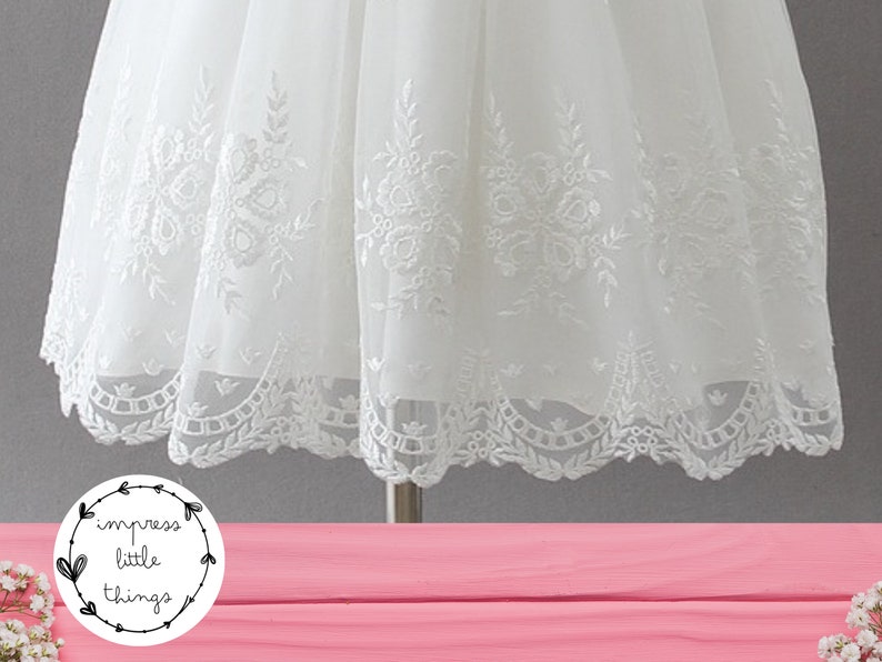 May include: Close-up of a white dress with intricate floral embroidery. The dress features a delicate, layered design with detailed patterns along the hemline. The image includes the text "impress little things" in a circular logo.