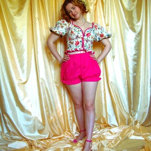 Puede incluir: Un top con estampado floral con mangas abullonadas y ribete rosa, combinado con shorts rosa brillante. El conjunto se completa con sandalias rosas. El fondo es una cortina de color amarillo dorado.