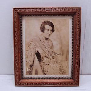 Puede incluir: Fotografía enmarcada en sepia de una mujer en un marco decorativo marrón. La mujer lleva un chal y tiene el pelo corto. El marco tiene un patrón detallado.