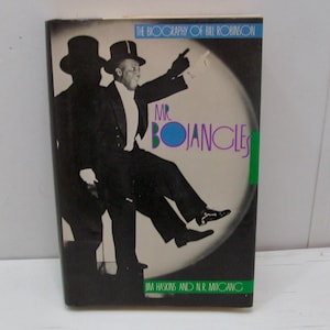 May include: A book cover featuring a black and white image of a man in a tuxedo and top hat, with the title "Mr. Bojangles" in purple and green. The book is a biography of Bill Robinson.