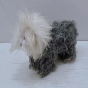 May include: A soft toy dog with a shaggy, two-tone coat. The dog has a fluffy white head and a grey body. The toy is standing on a white surface.
