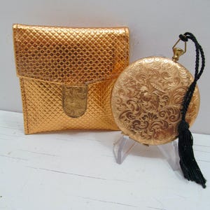 May include: A gold-toned pocket watch with intricate floral engravings and a black tassel, next to a matching gold pouch. The watch is round and ornate, while the pouch has a textured, grid-like pattern.