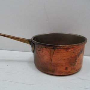May include: A copper saucepan with a long, brass-colored handle. The pot has a rounded shape and a tarnished, aged appearance. The interior is a lighter metal color. The pan sits on a white surface.