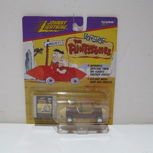 May include: A die-cast metal toy car of the Flintstones' car, a brown and cream vehicle with a yellow windshield, in a plastic blister pack. The packaging features a cartoon image of Fred and Barney Flintstone driving the car, along with the text "Cartoon Network The Flintstones".