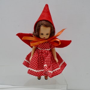 Puede incluir: Una muñeca pequeña con cabello castaño que lleva una capucha puntiaguda de fieltro rojo y un vestido rojo con lunares blancos. El vestido presenta un borde en zigzag blanco a lo largo del dobladillo. Una cinta naranja está atada en un lazo alrededor del cuello, y la muñeca tiene rasgos faciales pintados, incluyendo ojos oscuros y labios rojos.