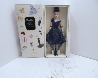 Puede incluir: Una muñeca Barbie de la colección Fashion Model Collection con un vestido de lunares negros y blancos, un sombrero negro y guantes blancos. La muñeca está en una caja blanca con un diseño rosa y blanco.